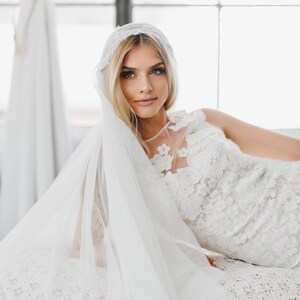 May include: A bride wearing a white lace wedding dress with a veil. The dress has a floral pattern and the veil is sheer and flowing.