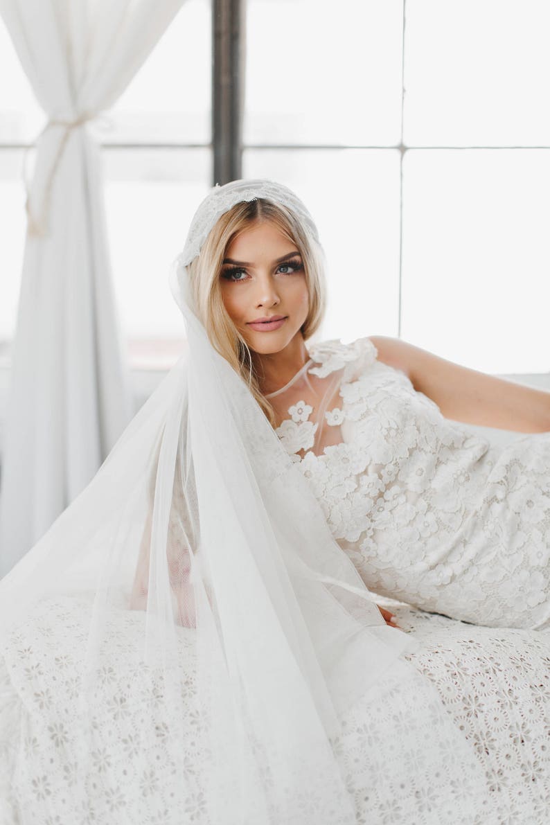 May include: A bride wearing a white lace wedding dress with a veil. The dress has a floral pattern and the veil is sheer and flowing.