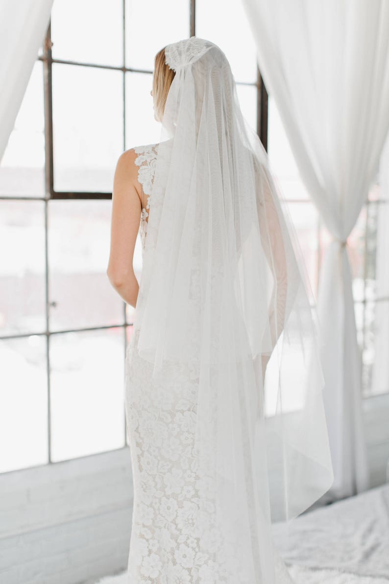 May include: A bride wearing a white lace wedding dress with a long, flowing veil. The veil is made of tulle and has a delicate lace trim. The bride is standing in front of a window, looking out at the view.