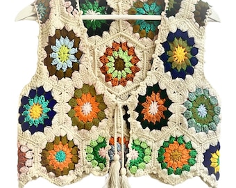 Puede incluir: Un chaleco de ganchillo con cierre de corbata y borla. El chaleco está hecho de hilo beige y presenta motivos florales coloridos en tonos de verde, azul, naranja y rosa.