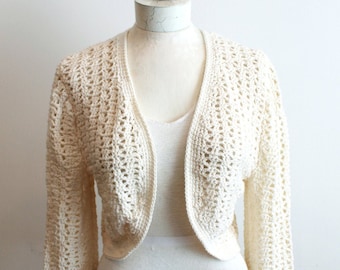 May include: A white crocheted cardigan with long sleeves and a V-neck. The cardigan is cropped and has a delicate openwork pattern.