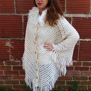 May include: White crochet poncho with a fringe border and a faux fur collar. The poncho has a button closure.