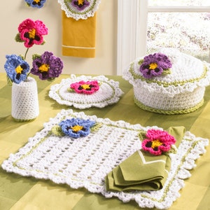 May include: A set of crocheted pansy flower themed kitchen accessories, including a pot holder, a bread basket cover, a set of placemats, and a vase. The items are all white with green accents and feature purple, pink, and yellow pansy flowers.