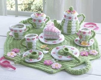 May include: A green crocheted play mat with a handle, featuring a tea set with a teapot, cups, saucers, and a cake. The set is decorated with pink and white flowers.