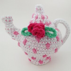 May include: A crocheted teapot with a pink and white polka dot pattern. The teapot has a green crocheted bow with a pink crocheted flower on top.