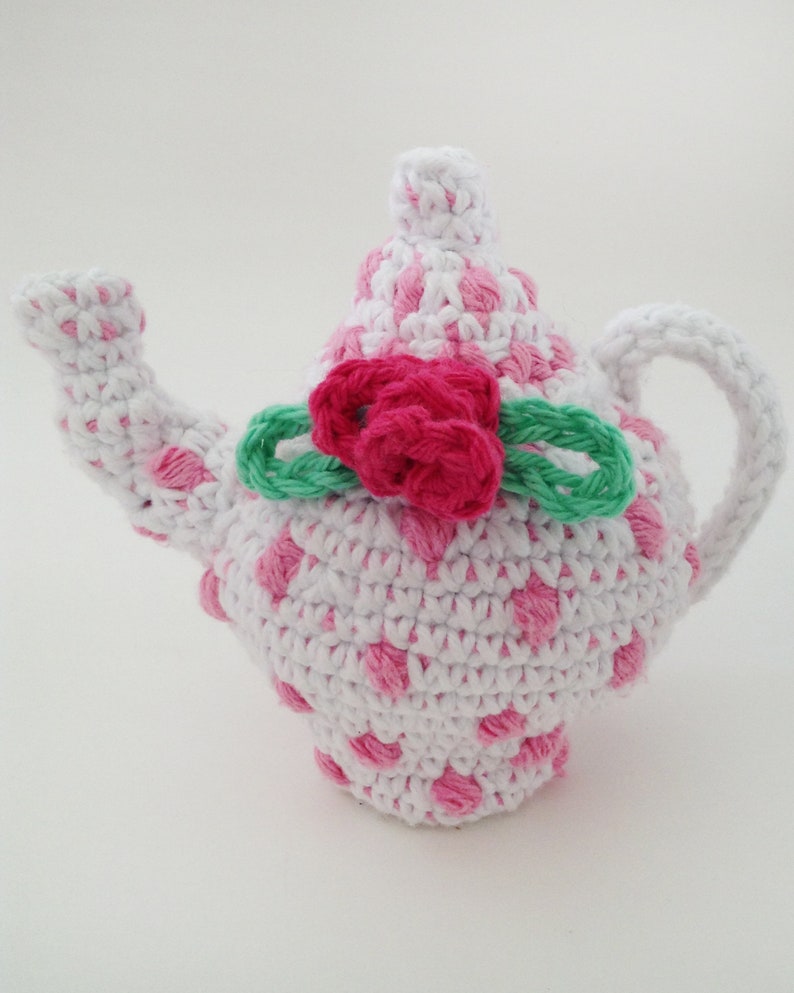 May include: A crocheted teapot with a pink and white polka dot pattern. The teapot has a green crocheted bow with a pink crocheted flower on top.