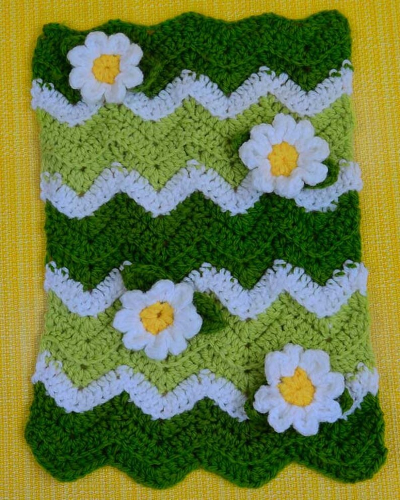 May include: A handmade crochet blanket featuring a green and white chevron pattern with white daisy accents. The blanket has a scalloped edge and is made with various shades of green yarn. The daisies have yellow centers.