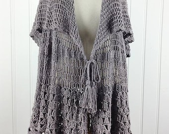 Könnte beinhalten: Ein grauer Häkelcardigan mit einem Bindeband vorne und einem Quaste. Der Cardigan hat eine lockere Passform und einen gewellten Saum.