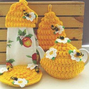 Puede incluir: Un conjunto de calienta tazas, calienta tetera y calienta taza de té de ganchillo en amarillo con flores blancas y detalles de abejas. El conjunto está hecho para parecerse a un panal.