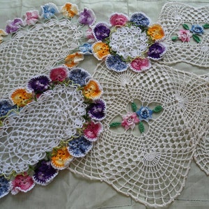 Könnte beinhalten: Vier weiße Häkeldeckchen mit Blumenmuster. Die Deckchen sind mit bunten Blumen verziert, darunter lila, rosa, blau und gelb.