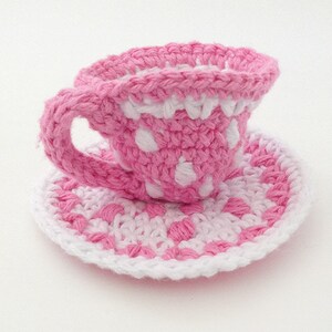 May include: A pink and white crocheted teacup and saucer set with a polka dot pattern.