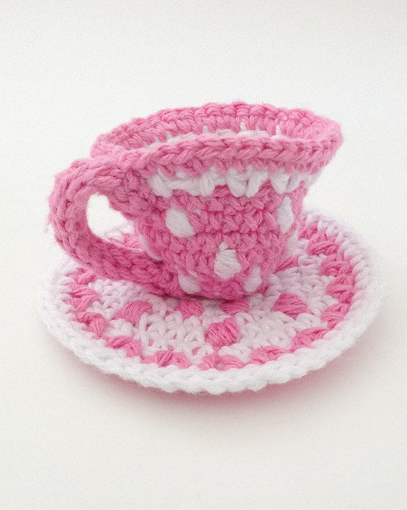 May include: A pink and white crocheted teacup and saucer set with a polka dot pattern.