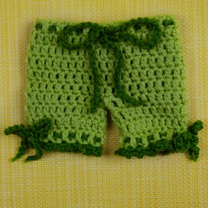 May include: Handmade green crochet shorts with a dark green drawstring and decorative trim. The shorts are made of a light green yarn and feature a scalloped edge. The shorts are displayed on a yellow textured background.