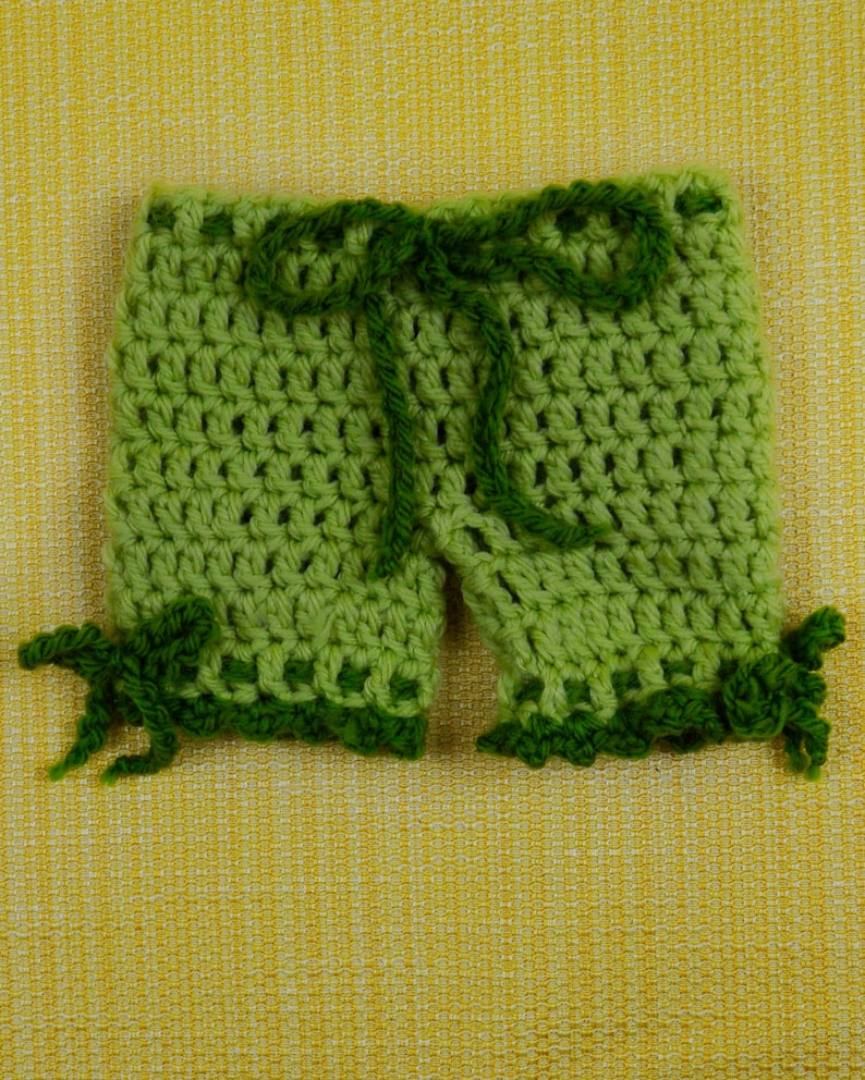 May include: Handmade green crochet shorts with a dark green drawstring and decorative trim. The shorts are made of a light green yarn and feature a scalloped edge. The shorts are displayed on a yellow textured background.