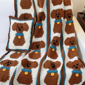 May include: A crocheted blanket and pillow featuring a brown dog design. The blanket is white with brown dogs and blue and white borders. The pillow is brown with a brown dog and blue and white borders.