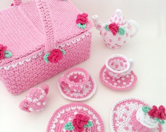 May include: A pink crocheted tea set with a teapot, cups, saucers, and a cupcake. The set is decorated with white polka dots and pink roses.