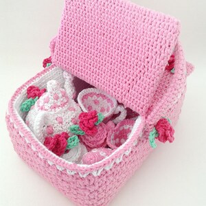 May include: A pink crocheted tea set storage basket with a lid. The basket is filled with a crocheted teapot, cups, saucers, and a sugar bowl. The basket is decorated with pink roses.