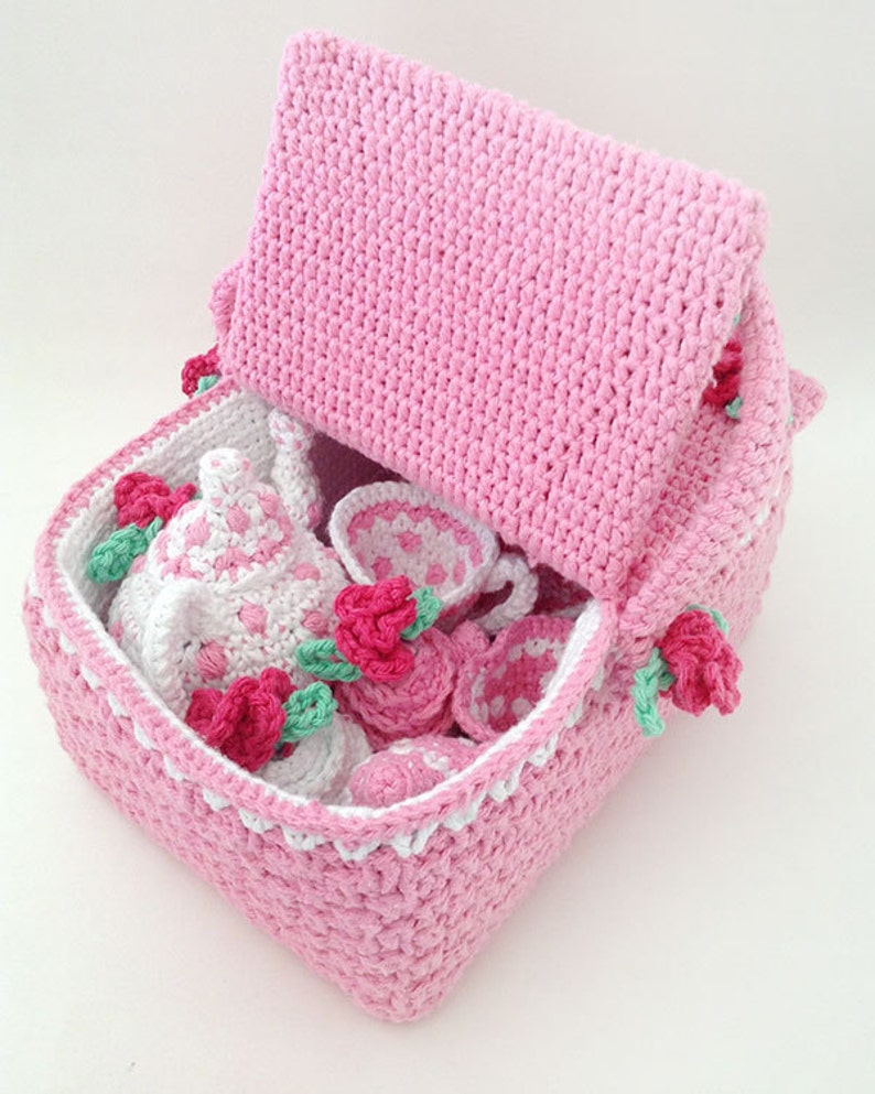 May include: A pink crocheted tea set storage basket with a lid. The basket is filled with a crocheted teapot, cups, saucers, and a sugar bowl. The basket is decorated with pink roses.