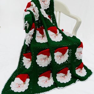 May include: A green crocheted blanket with a white border and multiple Santa Claus faces. The blanket is folded and draped over a white chair.