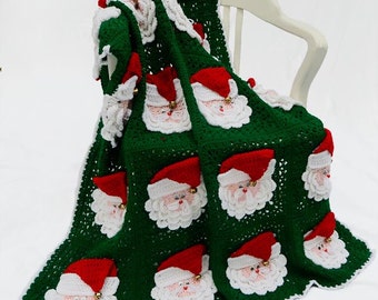 May include: A green crocheted blanket with a white border and multiple Santa Claus faces. The blanket is folded and draped over a white chair.