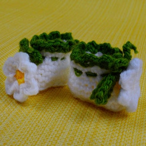May include: A pair of handmade, white crochet baby booties with green trim and accents. One bootie features a white flower with a yellow center. The booties are set against a yellow background.