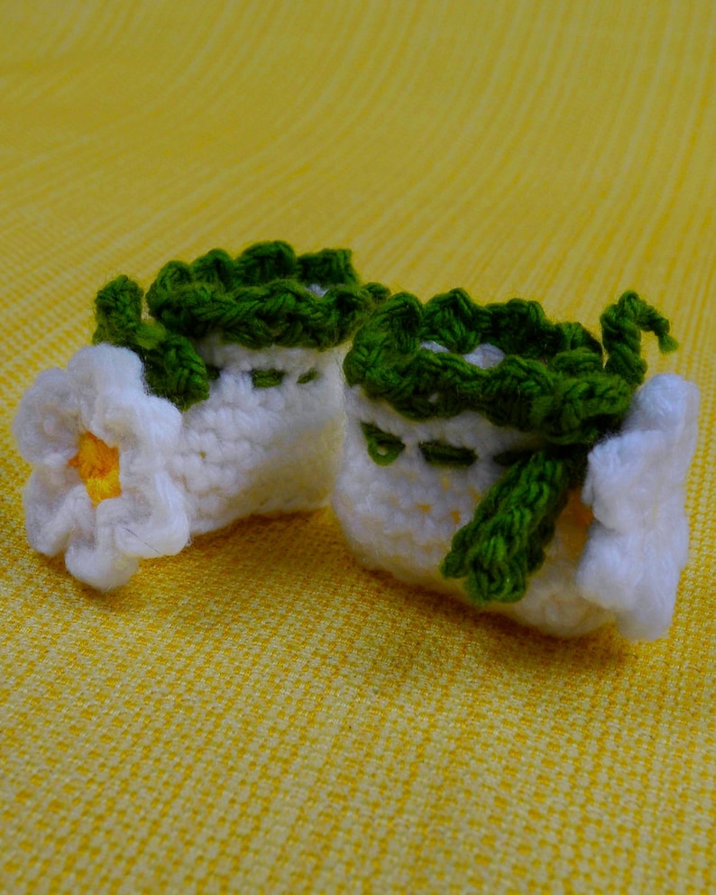 May include: A pair of handmade, white crochet baby booties with green trim and accents. One bootie features a white flower with a yellow center. The booties are set against a yellow background.