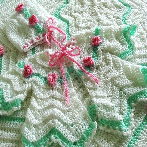 May include: A white crocheted baby blanket with a green border and pink flower accents.