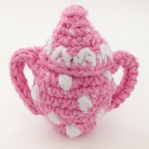 May include: A pink crocheted teapot with white polka dots. The teapot has a lid and two handles.