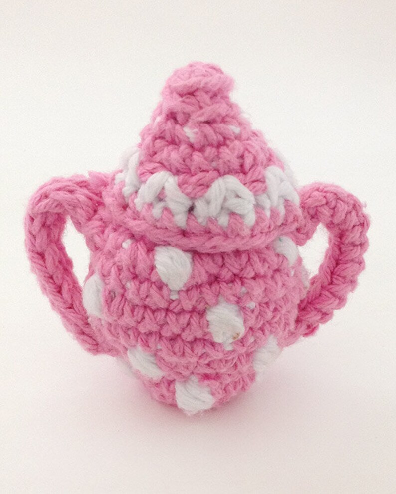 May include: A pink crocheted teapot with white polka dots. The teapot has a lid and two handles.