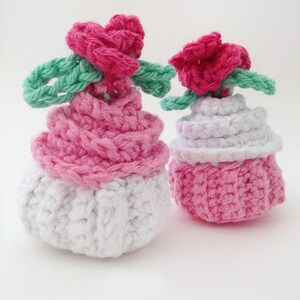 May include: Two crocheted cupcakes, one pink and white and the other all pink, with pink flower decorations on top.