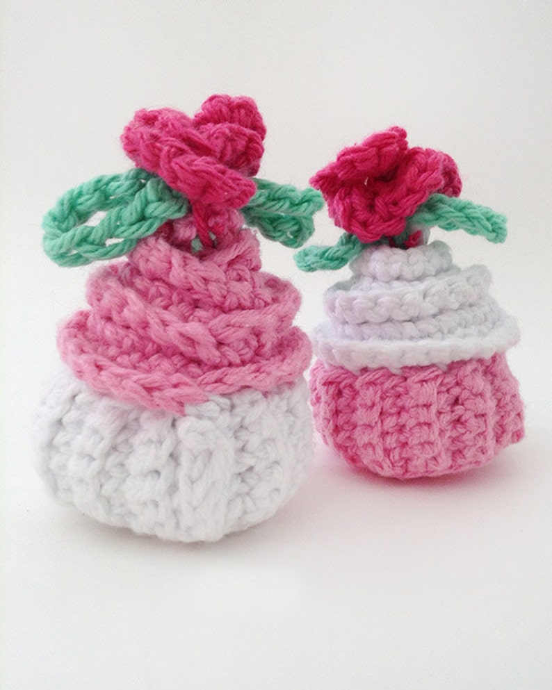 May include: Two crocheted cupcakes, one pink and white and the other all pink, with pink flower decorations on top.