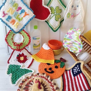 May include: A collection of colorful crocheted dishcloths with various holiday themes, including a heart, a shamrock, a bunny, a pumpkin, a turkey, a Christmas tree, a flower, a hat, and an American flag. The dishcloths are hanging on a white wooden wall with clothespins.