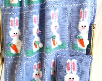 Pode incluir: Um cobertor e uma almofada para bebé em crochê com um padrão repetido de coelhos brancos segurando cenouras laranja. O cobertor é azul claro com detalhes verdes e brancos. A almofada tem o mesmo design que o cobertor.