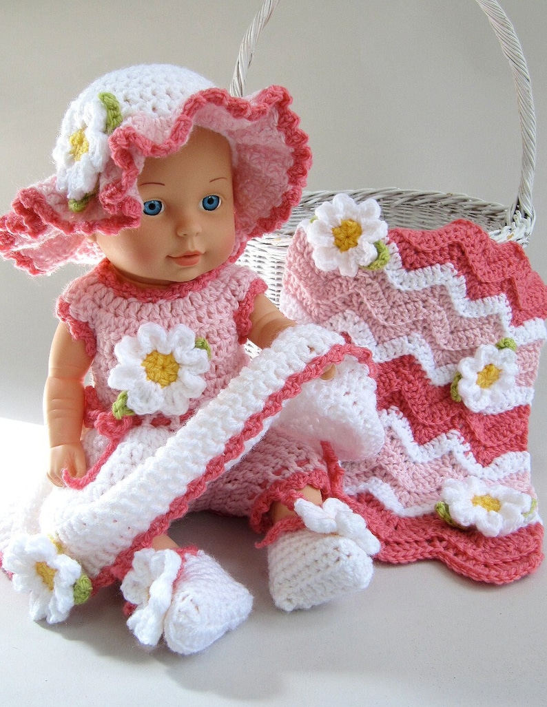 May include: A crochet doll wearing a pink and white outfit with daisy accents. The outfit includes a hat, dress, shoes, and a blanket. The doll has blue eyes and is sitting next to a white basket.