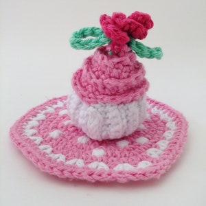 May include: A pink and white crocheted cupcake with a pink flower on top, sitting on a pink and white crocheted doily.