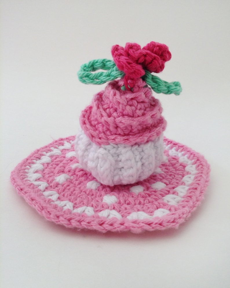 May include: A pink and white crocheted cupcake with a pink flower on top, sitting on a pink and white crocheted doily.