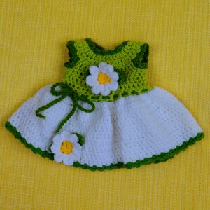 May include: A handmade crochet dress in white, green, and yellow, featuring a daisy design. The bodice is green, and the skirt is white, with green trim. Two daisy flowers are attached to the dress. The dress is displayed on a yellow background.