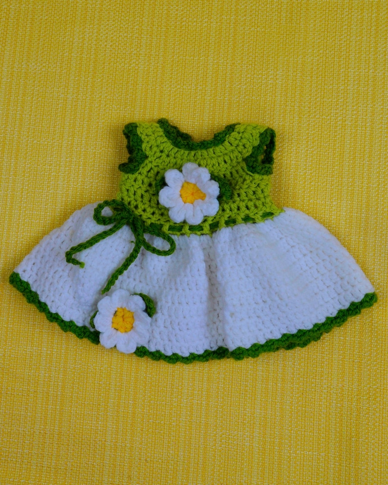 May include: A handmade crochet dress in white, green, and yellow, featuring a daisy design. The bodice is green, and the skirt is white, with green trim. Two daisy flowers are attached to the dress. The dress is displayed on a yellow background.