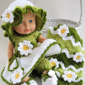 May include: A doll wearing a handmade crochet outfit, including a hat, dress, and shoes, all adorned with white and yellow daisy accents. The outfit is primarily white and green, with a matching blanket. A white wicker basket is in the background.