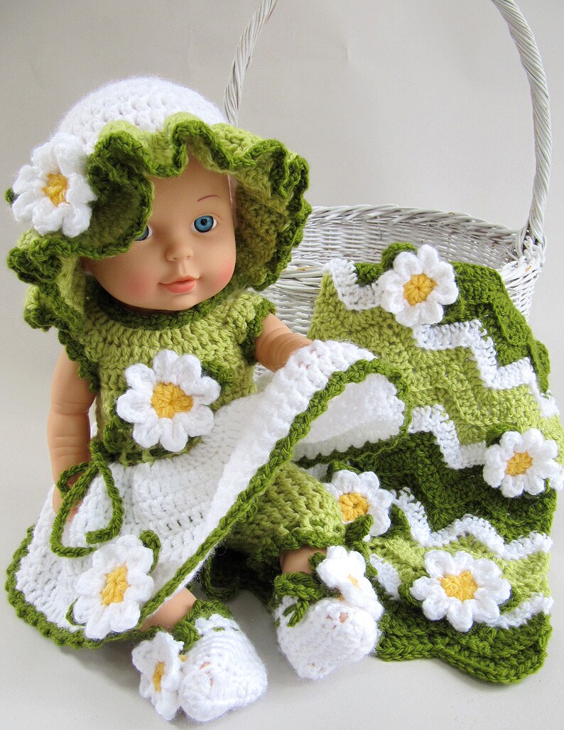 May include: A doll wearing a handmade crochet outfit, including a hat, dress, and shoes, all adorned with white and yellow daisy accents. The outfit is primarily white and green, with a matching blanket. A white wicker basket is in the background.