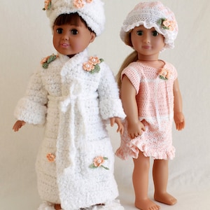 May include: Two dolls wearing crocheted outfits. The doll on the left is wearing a white robe with floral accents and white slippers. The doll on the right is wearing a pink dress with white trim and a white crocheted hat with a flower accent.
