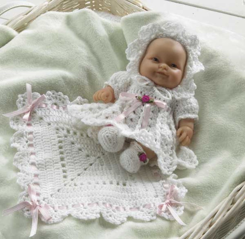 May include: A white crocheted baby doll dress with pink ribbon accents and a matching white crocheted blanket with pink ribbon trim. The doll is wearing a white crocheted bonnet.
