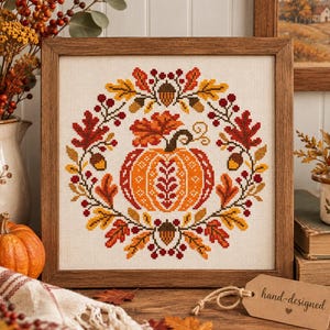 May include: A square framed cross-stitch embroidery featuring a central orange pumpkin surrounded by a wreath of autumn leaves, acorns, and red berries. The design is stitched on white fabric and enclosed in a medium-toned wooden frame.