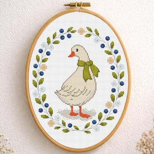 Könnte beinhalten: Ovale Kreuzstich-Stickerei mit einer weißen Gans mit orangefarbenem Schnabel und Füßen, die einen grünen Schal trägt. Die Gans ist von einem Kranz aus blauen Beeren, grünen Blättern und kleinen weißen und rosa Blüten umgeben, in einem Holzreifen.