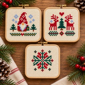 May include: Three square wooden embroidery hoops containing cross-stitch patterns on cream fabric. One features a gnome with a red hat, one shows two red reindeer flanking a green tree, and the third displays a red and green snowflake design.