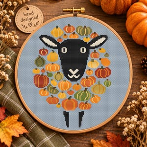 May include: A cross-stitch embroidery in a round wooden hoop featuring a black sheep head centred on a light blue background. The sheep's wool is depicted as a collection of various orange, yellow, and green pumpkins of different sizes.