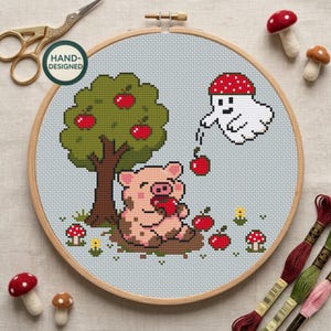 May include: A cross-stitch project in a wooden hoop featuring a pixelated pig eating an apple under a tree, a ghost holding an apple, and small mushrooms. The design includes a green tree with red apples, a white ghost, and a pink pig on a light blue background. Gold scissors and three felt mushrooms are nearby.