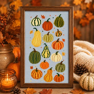 Puede incluir: Un bordado en punto de cruz enmarcado que presenta una colección de varias calabazas y calabacines en tonos naranja, verde, amarillo y blanco, dispuestos sobre un fondo de cuadrícula azul claro con pequeños motivos de hojas de otoño.