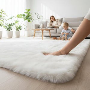 Op de afbeelding: Een groot, pluizig wit tapijt met een zacht, getextureerd oppervlak. Het tapijt is rechthoekig en bedekt een groot deel van de lichtgekleurde houten vloer. De voet van een persoon raakt het tapijt. De achtergrond toont een woonkamer met een bank en planten.