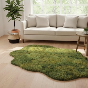 May include: A green, irregularly shaped rug with a moss-like texture. The rug is placed on a light wood floor in front of a cream-colored sofa. A potted plant and a small side table are also visible.